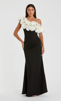 Woman in the Mac Duggal 21079 one-shoulder black prom gown, featuring large white floral appliqués across the top, stands against a plain white background.