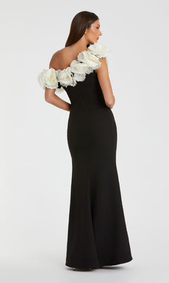 A woman stands against a plain background wearing the Mac Duggal 21079 one-shoulder black prom gown, featuring large white floral accents along the neckline.