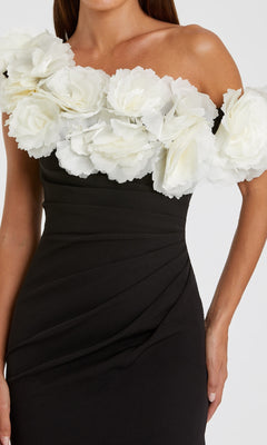 A woman wears the One-Shoulder Black Prom Gown by Mac Duggal 21079, featuring large white fabric flowers along the neckline and shoulders.