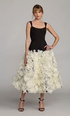 A woman wears the Mac Duggal 21080 midi wedding guest dress—black, sleeveless crepe with a voluminous skirt featuring large 3D organza flowers—paired with black high-heeled sandals.