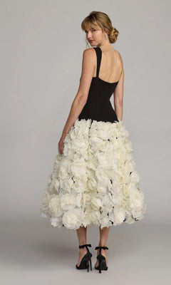 A woman stands in front of a plain backdrop, wearing the Mac Duggal 21080 Midi Wedding Guest Dress—a sleeveless black crepe design with a voluminous skirt embellished with 3D organza flowers—glancing over her shoulder.