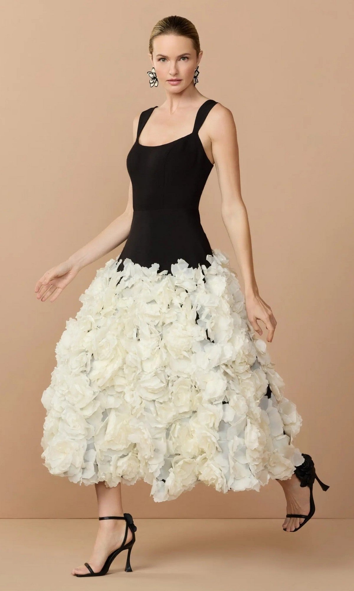 A woman wears the Mac Duggal 21080 Midi Wedding Guest Dress in Black Cream, featuring a sleeveless fitted bodice and a voluminous cream skirt with 3D organza flowers, paired with black heels, against a beige background.