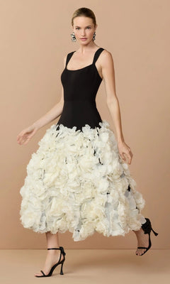 A woman wears the Mac Duggal 21080 Midi Wedding Guest Dress in Black Cream, featuring a sleeveless fitted bodice and a voluminous cream skirt with 3D organza flowers, paired with black heels, against a beige background.