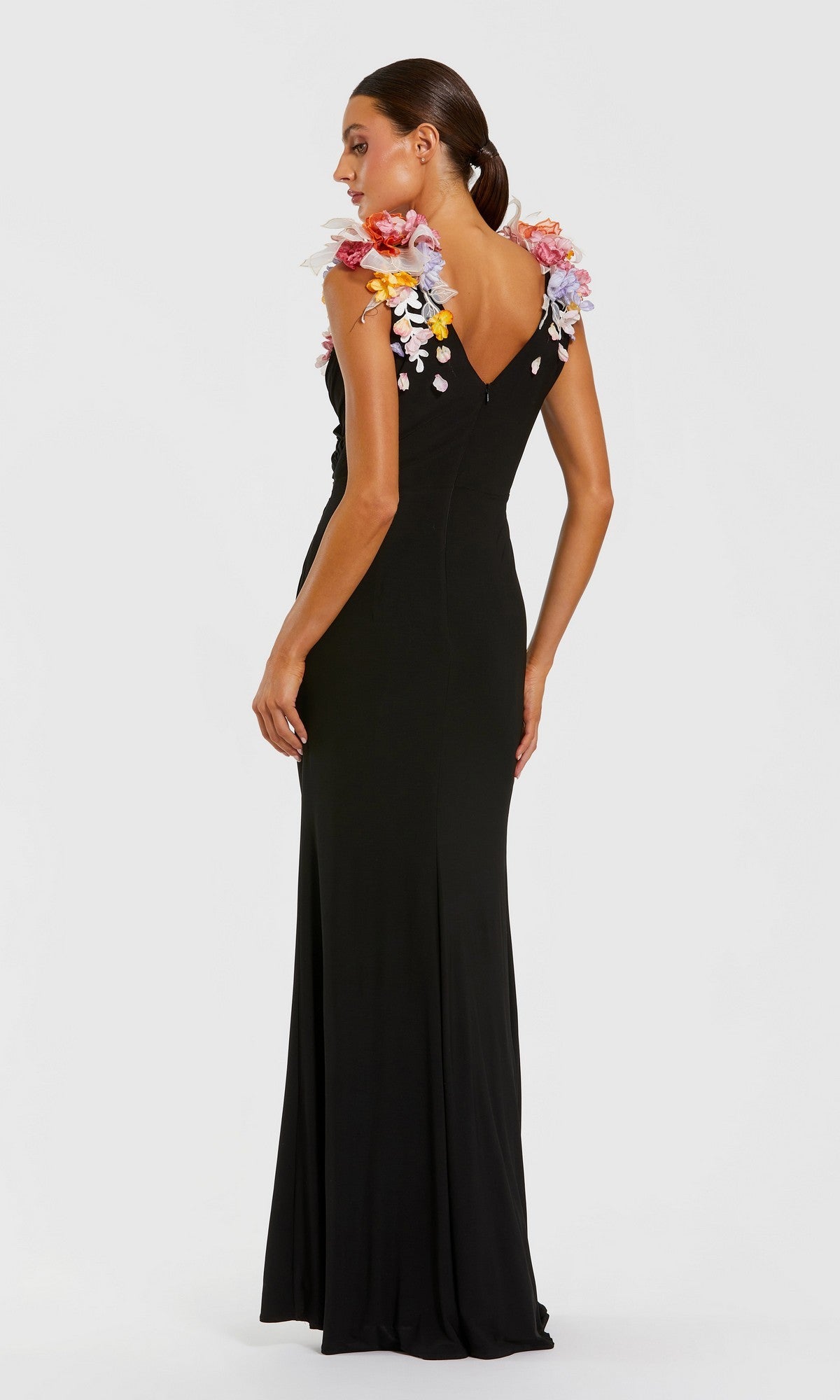 A woman stands facing slightly away from the camera against a plain background, wearing the Mac Duggal 21084 long formal dress in black, which features vibrant 3D floral appliques on the shoulders.