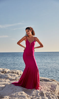 A woman in the Deep V-Neck Long Prom Dress: Aerie Couture 210, a fitted magenta gown with thin straps and a lace-up open back, stands on a rocky shore by the ocean under a clear sky.