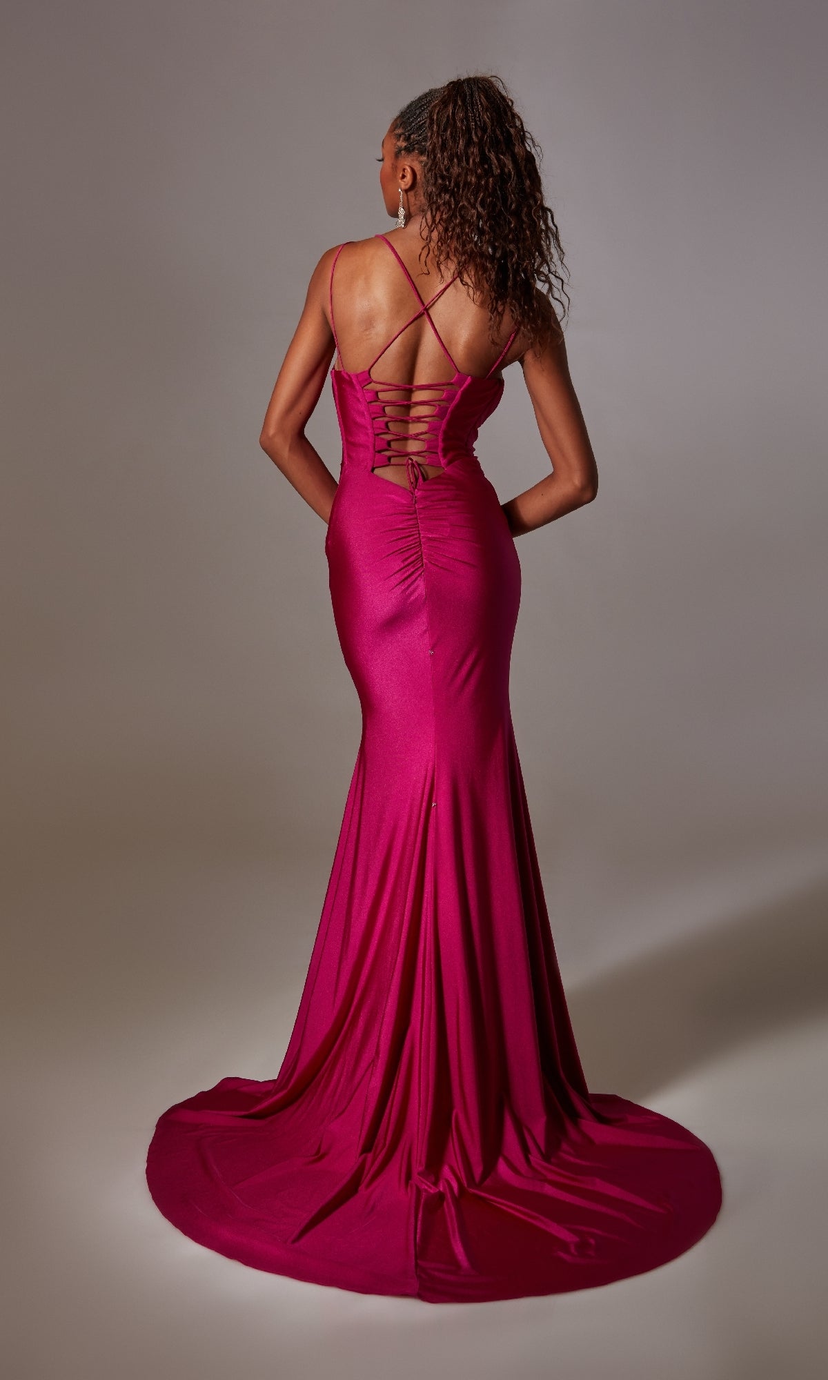 A woman models the Deep V-Neck Long Prom Dress: Aerie Couture 210, a magenta gown with a fitted silhouette and crisscross lace-up open back, standing before a plain gray background.