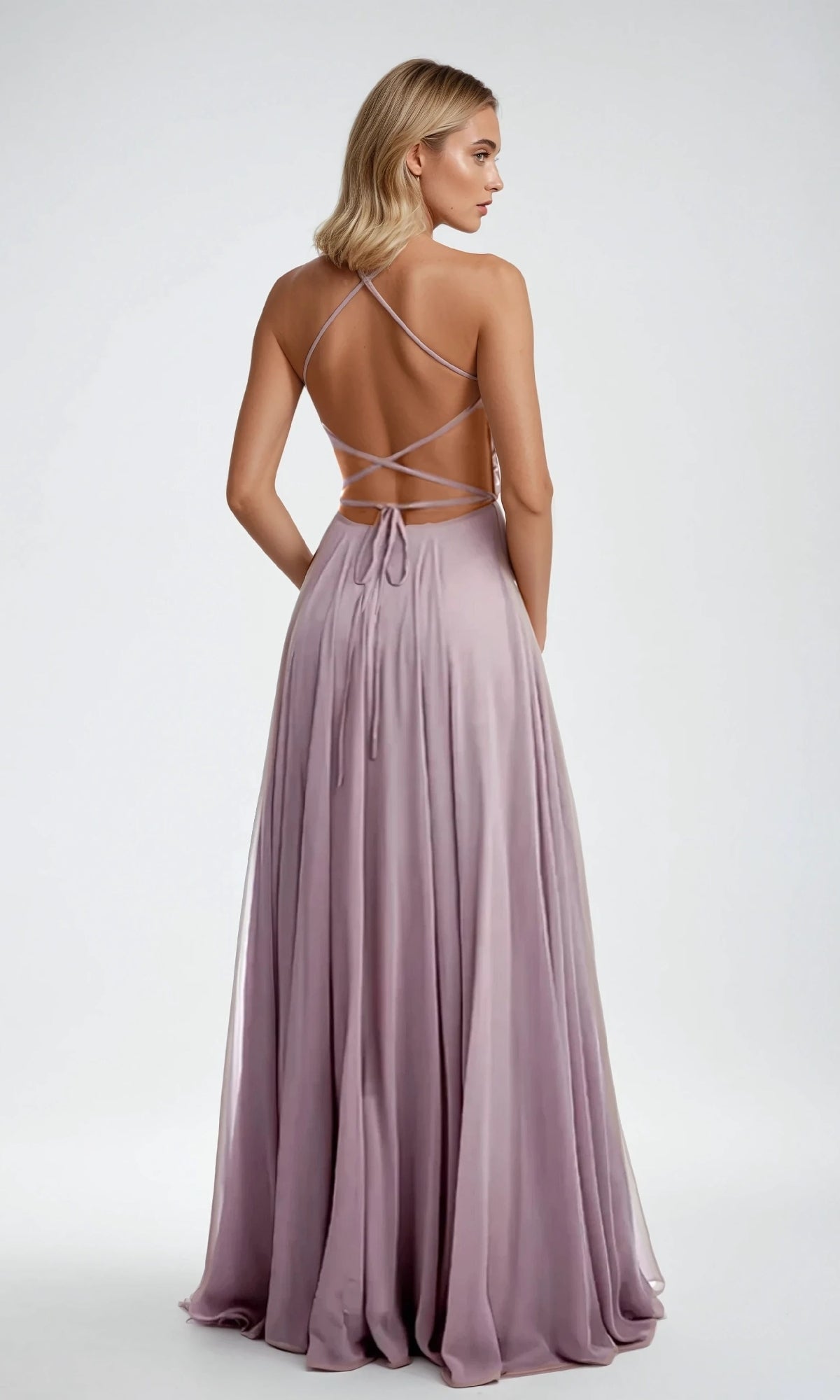 A woman wearing the Long Prom Dress: Juliet 210, featuring a lavender backless design with crisscross straps and an elegant A-line skirt, stands facing away from the camera against a plain background.