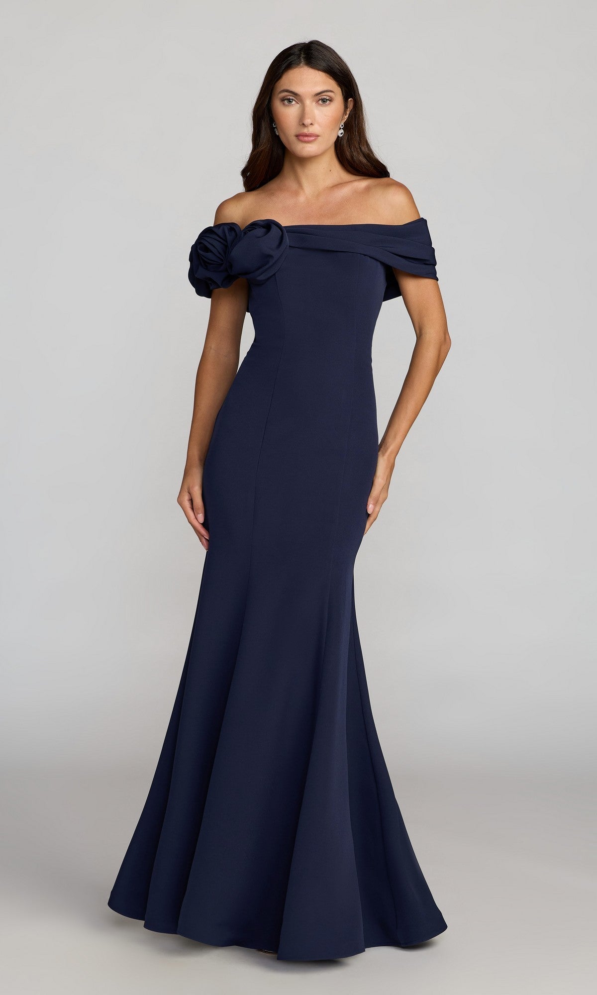 A woman models the Off-Shoulder Long Formal Dress: Mac Duggal 21104, a floor-length mermaid gown featuring a large fabric flower accent, standing against a plain background to highlight its elegant design.