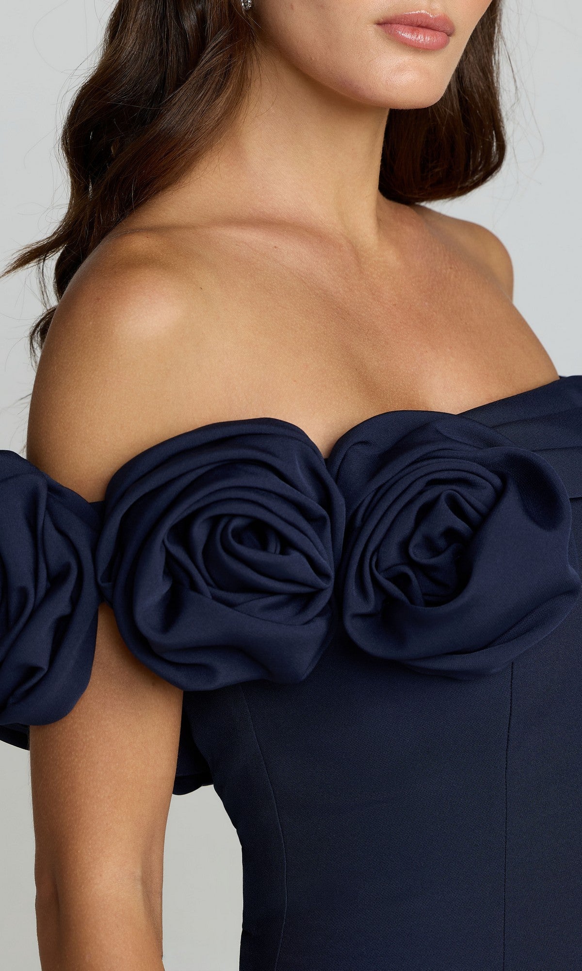 A woman in the Mac Duggal 21104 off-shoulder long formal dress in navy, featuring a mermaid silhouette and large fabric roses along the neckline.