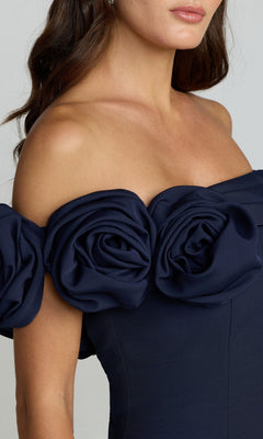 A woman in the Mac Duggal 21104 off-shoulder long formal dress in navy, featuring a mermaid silhouette and large fabric roses along the neckline.