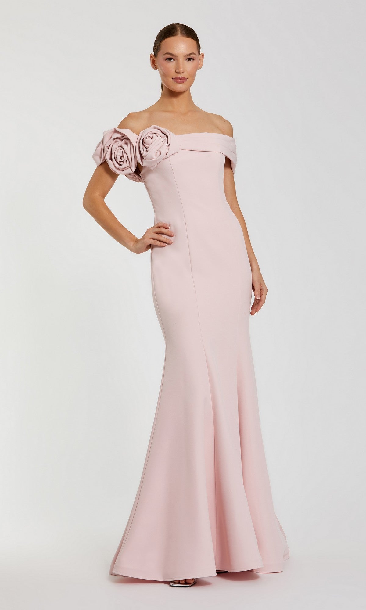 A woman stands with one hand on her hip, modeling the Mac Duggal 21104: a pale pink off-shoulder mermaid formal dress featuring two large fabric roses on one sleeve, set against a plain background.