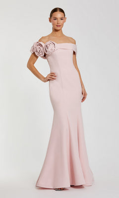 A woman stands with one hand on her hip, modeling the Mac Duggal 21104: a pale pink off-shoulder mermaid formal dress featuring two large fabric roses on one sleeve, set against a plain background.