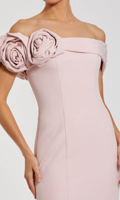 A woman wears the Mac Duggal 21104 Off-Shoulder Long Formal Dress, a light pink mermaid gown featuring large rose details along the neckline.