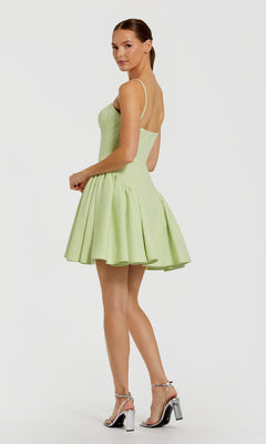 A woman poses in the Mac Duggal 21124 short wedding guest dress—light green, sleeveless, flared with spaghetti straps—and silver high-heeled sandals against a plain white background.
