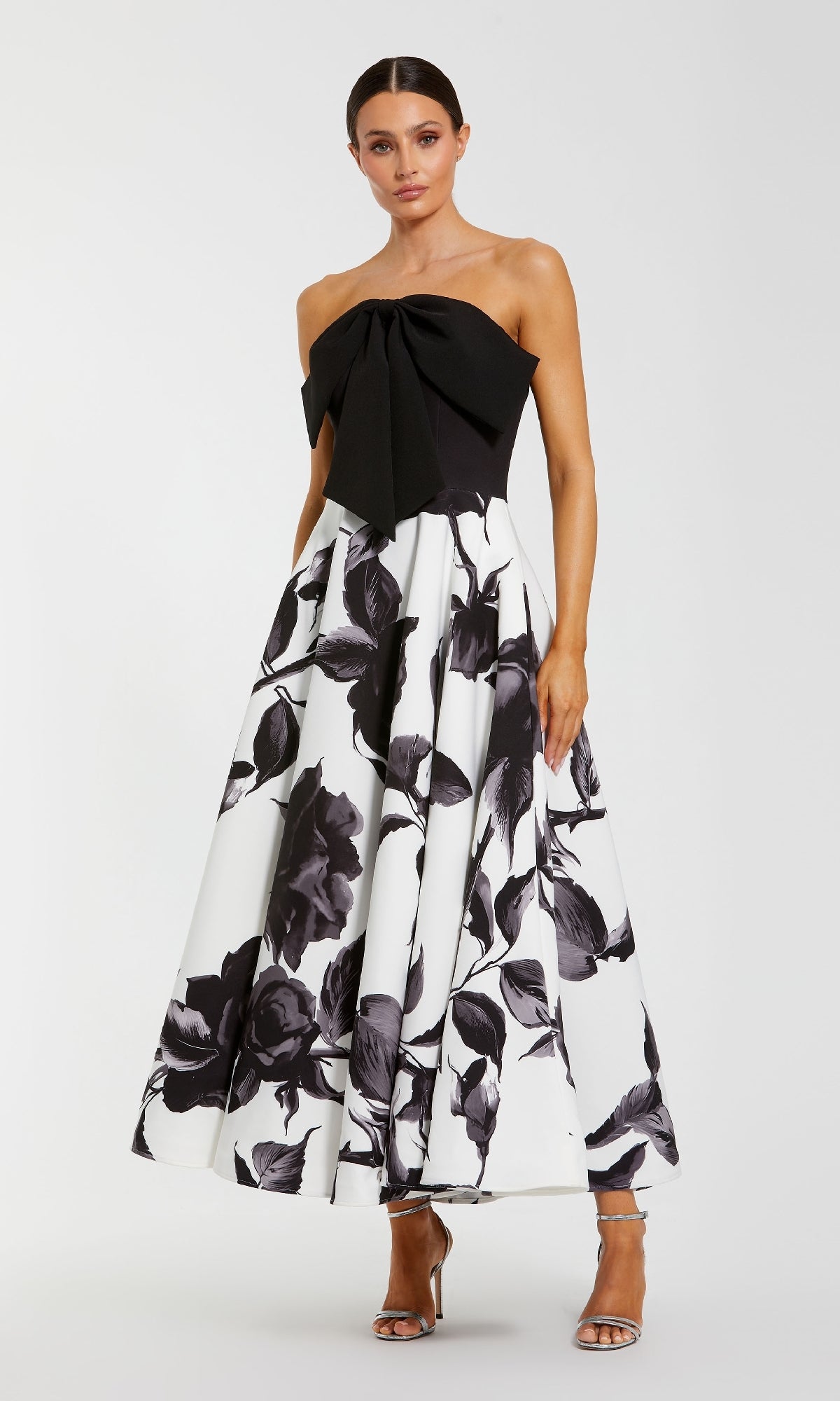 A woman models the Mac Duggal 21151 long formal dress, a strapless black and white floral gown with a large black bow on the bodice, set against a simple background.
