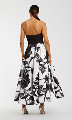 A woman stands with her back to the camera, wearing the Mac Duggal 21151 long formal dress— a strapless black and white floral evening gown—paired with silver high-heeled sandals.