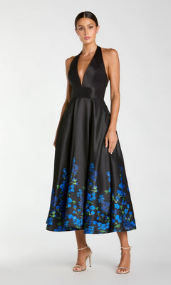 A woman wearing the Mac Duggal 21154 tea-length party dress in Black Multi, featuring a black satin halter-neck design with a deep V-neckline, sleeveless silhouette, and blue floral patterns near the hem, paired elegantly with strappy high-heeled sandals.