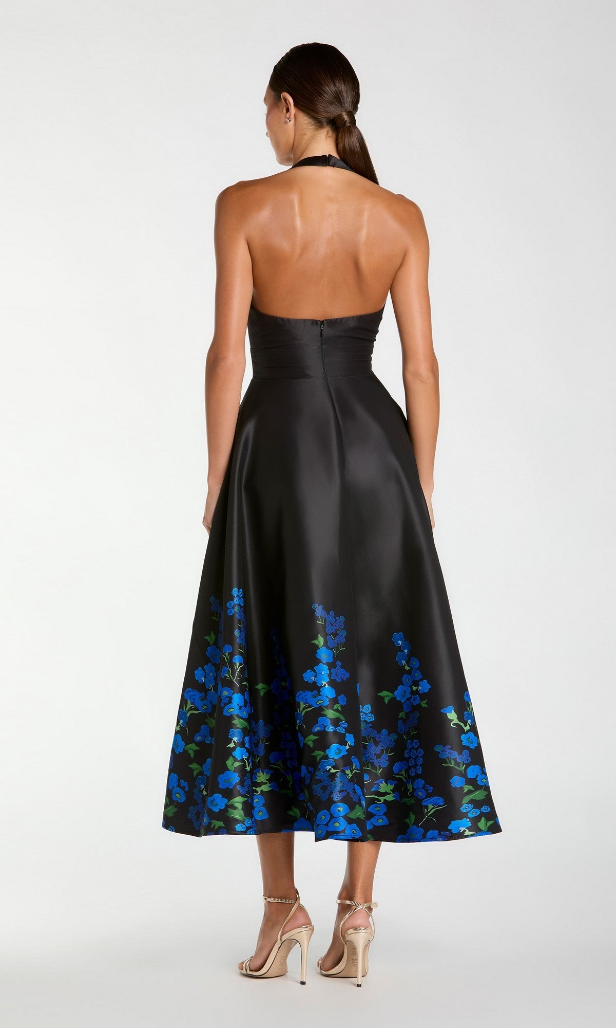 Wearing the Tea-Length Party Dress: Mac Duggal 21154—a strapless black midi dress with a blue floral print at the hem—she stands with her back to the camera, paired with beige high-heeled sandals.