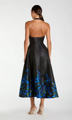 Wearing the Tea-Length Party Dress: Mac Duggal 21154—a strapless black midi dress with a blue floral print at the hem—she stands with her back to the camera, paired with beige high-heeled sandals.