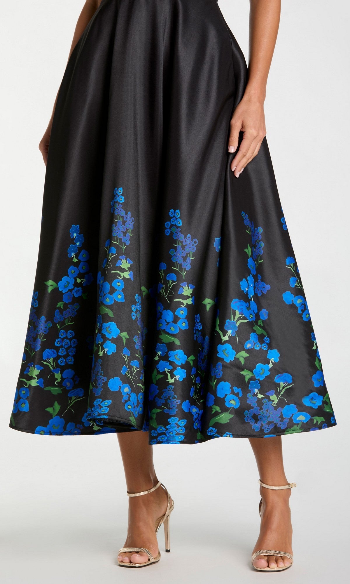 Wearing the Mac Duggal 21154 Tea-Length Party Dress—a black dress adorned with blue floral prints—paired with gold high-heeled sandals, a person stands against a plain background.