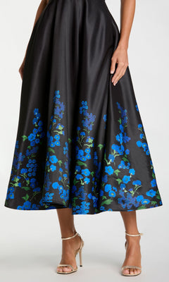 Wearing the Mac Duggal 21154 Tea-Length Party Dress—a black dress adorned with blue floral prints—paired with gold high-heeled sandals, a person stands against a plain background.