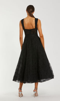 Woman stands with her back to the camera in the Mac Duggal 21171, a sleeveless, textured tea-length black formal dress, paired with silver high-heeled sandals against a plain background.