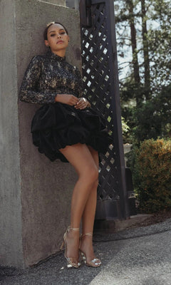 A woman in a black Mac Duggal 21175 short semi-formal dress with a lace mock-neck and ruffled skirt leans against an outdoor wall, wearing silver heels. Trees and bushes are in the background.