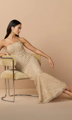 A woman wearing the Mac Duggal 21200 long formal dress in gold sits sideways on a beige chair against a beige background, looking to her left.
