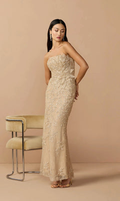 A woman wears the Mac Duggal 21200 long formal dress—a gold strapless gown with intricate beading—standing beside a light-colored chair against a plain beige background.