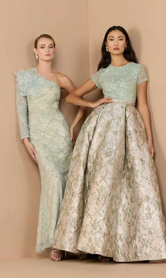 Two women pose against a beige backdrop in elegant light blue and silver gowns featuring intricate beadwork and floral designs—one dazzles in a sequin-beaded dress, while the other wears the graceful Mac Duggal 21203 long formal dress.