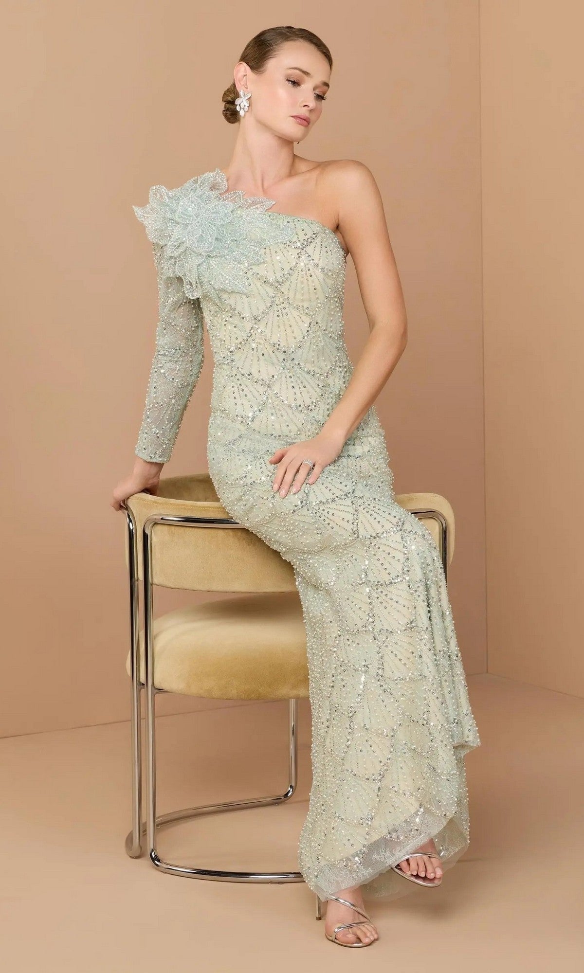 A woman in a seafoam Mac Duggal 21203 long formal one-shoulder gown with beading and floral appliqué sits on a beige chair against a plain tan background.
