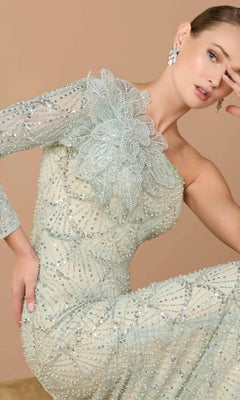 A woman wearing the Mac Duggal 21203 long formal dress—featuring light sequin beading, a one-shoulder design, and a large floral accent—poses with one arm bent near her face against a neutral background.