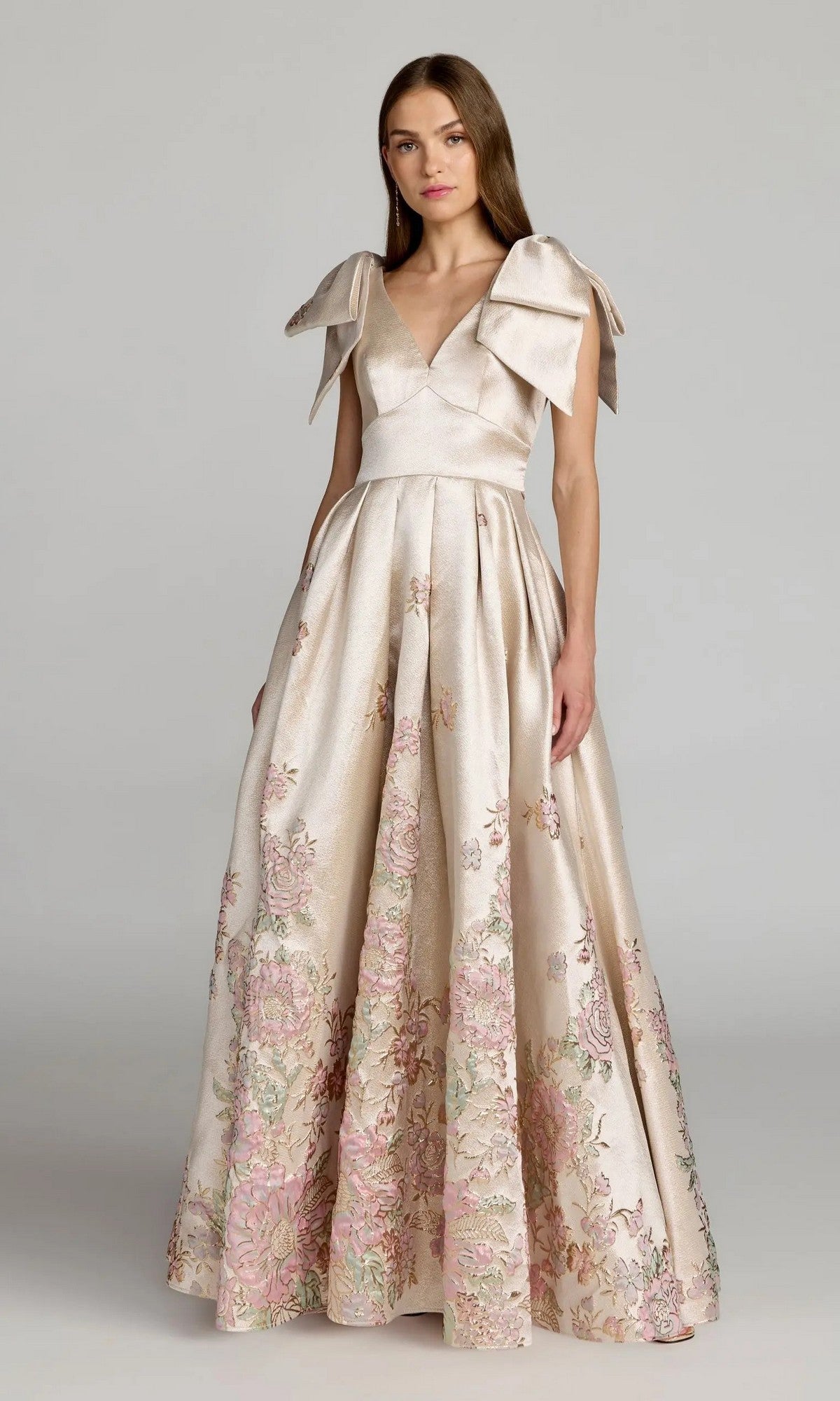 A woman stands against a plain background wearing the Mac Duggal 21204 long formal dress in Champagne Multi, featuring a floor-length satin brocade fabric with floral embroidery, cap sleeves adorned with bows, and a V-neckline in an elegant ballgown silhouette.