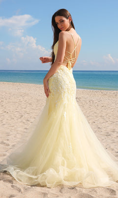 A woman in a pale yellow Amelia Couture 2120 long mermaid prom dress with an embroidered lace back stands on a sandy beach, facing the camera with the ocean and blue sky behind her.