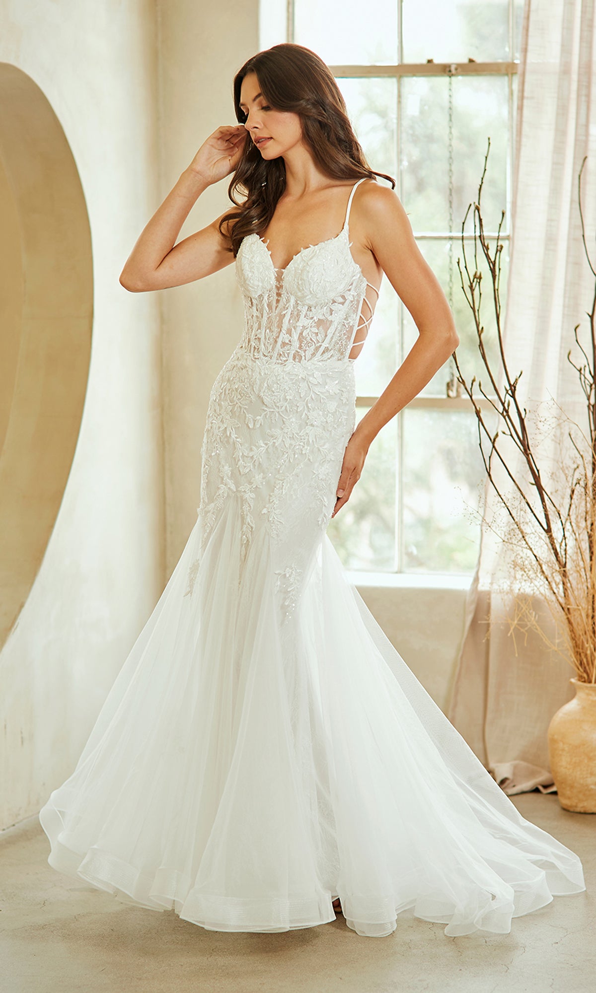 A woman poses indoors by a window in the Amelia Couture 2120 Long Mermaid Prom Dress, a fitted, sleeveless white embroidered lace gown with a flared tulle skirt.