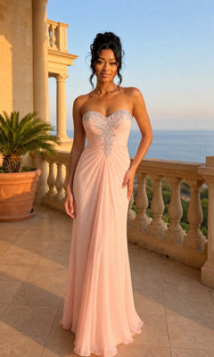 Wearing the La Femme Dress 21214 in apricot, a woman stands on a stone balcony overlooking the sea. The strapless chiffon gown features silver embellishments and a ruched bodice.