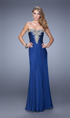 A woman models the La Femme Dress 21214, a royal blue strapless sweetheart gown with a floor-length skirt and silver-embellished ruched bodice, standing before a simple gray background.