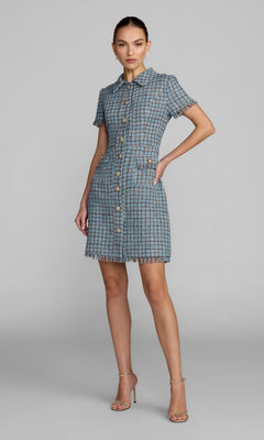 A woman stands with one hand on her hip, modeling the Mac Duggal 21221 Short Boucle Day Dress in Blue Multi. The mini dress features short sleeves, a collared neckline, frayed edges, and gold button detailing. She pairs the look with nude heels against a plain gray background.