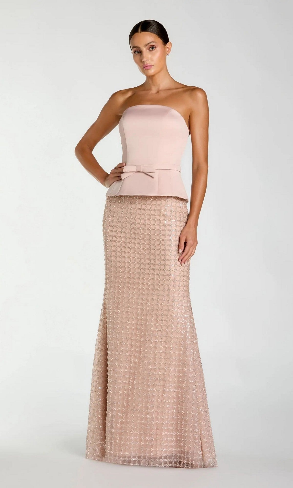 A woman stands against a plain light background wearing the Mac Duggal 21227 long formal dress in blush, featuring a strapless fitted bodice, peplum detail, and a textured floor-length skirt adorned with delicate beaded-lattice embellishments.