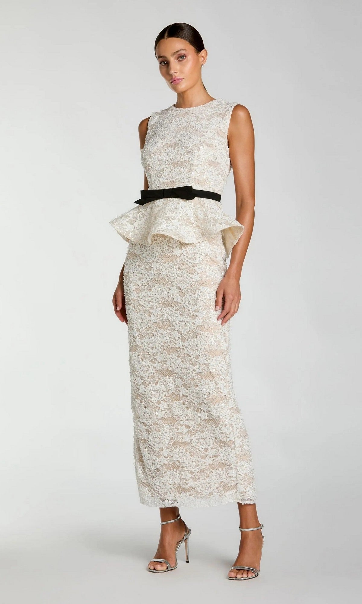 A woman stands against a plain background wearing the Mac Duggal 21228, a sleeveless, floor-length ivory nude lace formal dress featuring a peplum waist and black belt, paired with silver high-heeled sandals.