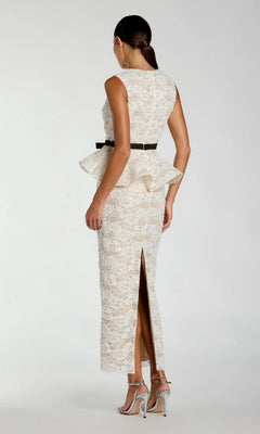 A woman stands in profile against a plain background, wearing the Mac Duggal 21228 long formal dress—an elegant ivory lace gown featuring a peplum waist, black belt, back slit, and silver high-heeled sandals.