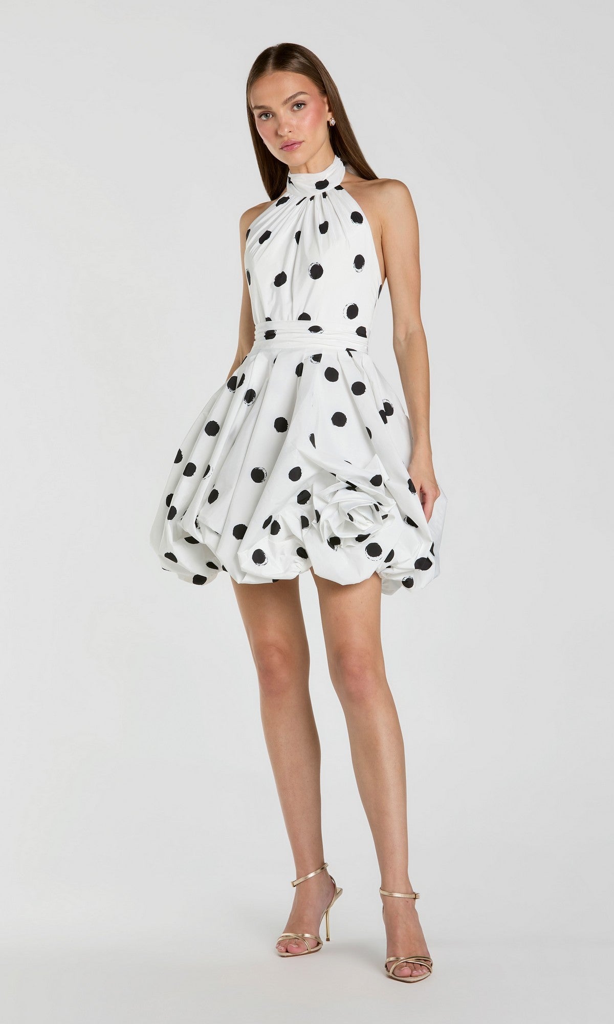 A woman wearing the Mac Duggal 21233 short polka dot party dress in black ivory, featuring a sleeveless halter-neck design, playful fit-and-flare silhouette, and bubble hemline, paired with strappy high-heeled sandals, stands against a plain background.