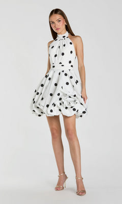A woman wearing the Mac Duggal 21233 short polka dot party dress in black ivory, featuring a sleeveless halter-neck design, playful fit-and-flare silhouette, and bubble hemline, paired with strappy high-heeled sandals, stands against a plain background.