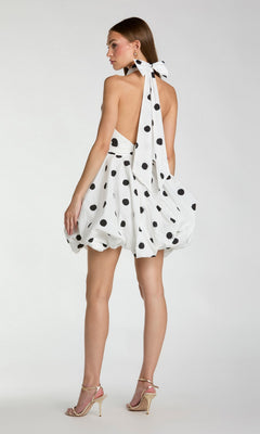 A woman models the Short Polka Dot Party Dress: Mac Duggal 21233—an eye-catching white dress with playful polka dots, a backless design, dramatic neck bow, and bubble hemline in a fit-and-flare silhouette—paired with high-heeled sandals, standing against a plain background.