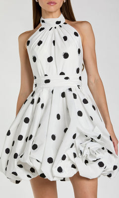 A woman wearing the Mac Duggal 21233 Short Polka Dot Party Dress, featuring a sleeveless, high-neck design with black polka dots on white fabric, a fitted waist, voluminous fit-and-flare silhouette, and a large fabric flower detail adorning the lower right side.