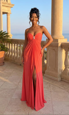 A woman stands on a stone terrace with ocean views, wearing La Femme Dress 21233 in Pink Grapefruit, featuring a strapless bodice and front slit.