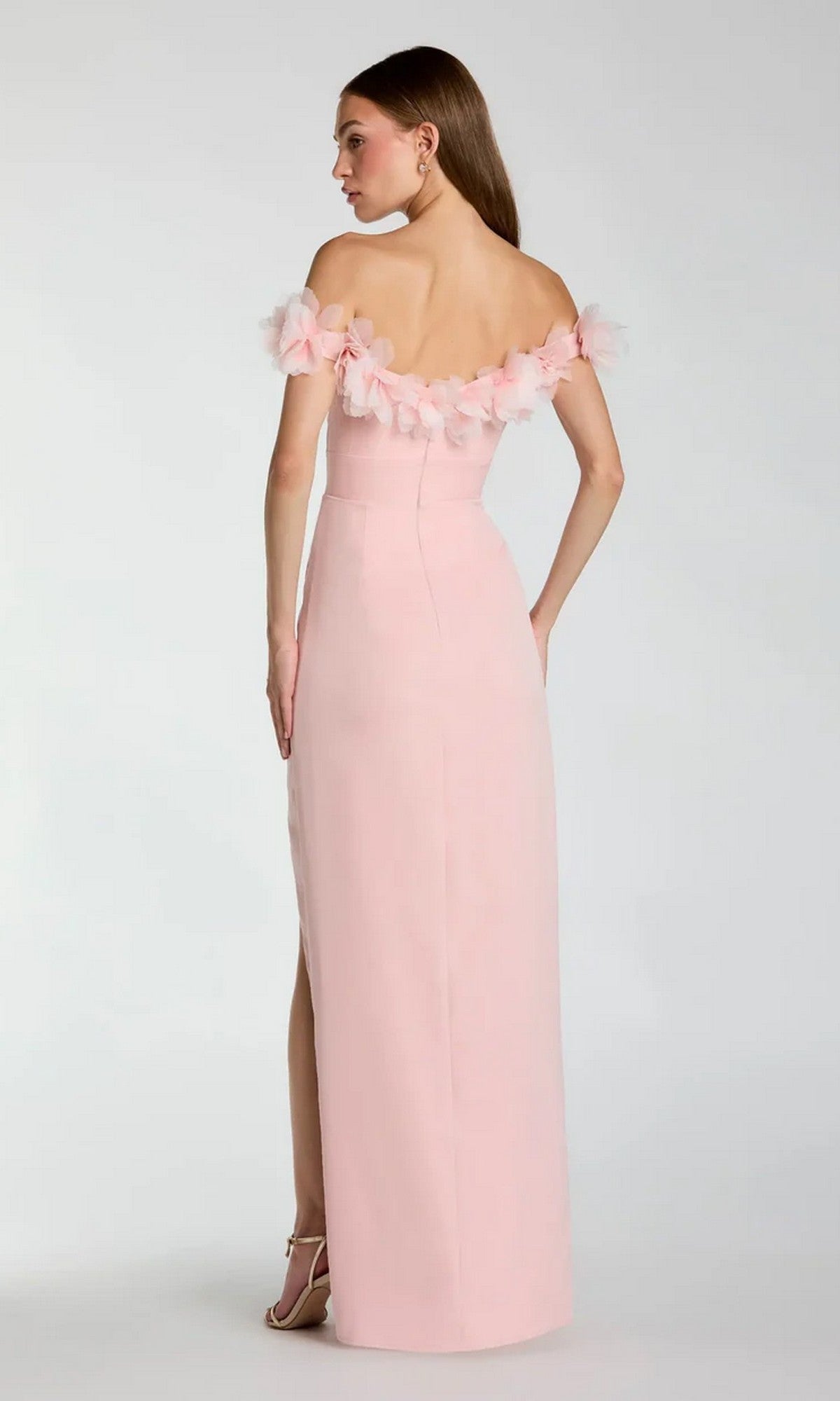 A woman stands facing away, wearing the Mac Duggal 21235 long formal dress in light pink crepe-jersey, featuring an off-shoulder sweetheart neckline with floral accents and a back slit, paired with strappy heels.