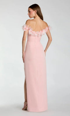 A woman stands facing away, wearing the Mac Duggal 21235 long formal dress in light pink crepe-jersey, featuring an off-shoulder sweetheart neckline with floral accents and a back slit, paired with strappy heels.