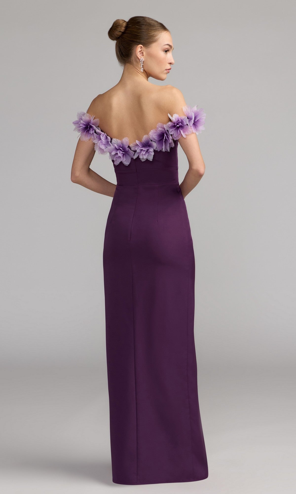 A woman stands facing away in a studio, wearing the Mac Duggal 21235 long formal dress—a floor-length, off-shoulder design in purple crepe-jersey with a sweetheart neckline and floral detailing along the neckline.