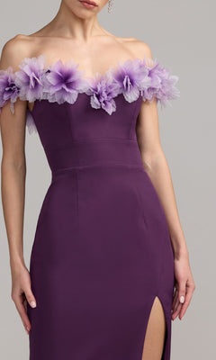 A woman wearing the Mac Duggal 21235 long formal dress—a purple off-the-shoulder gown with a sweetheart neckline, lilac floral accents along the neckline, and a thigh-high slit—stands against a neutral background.